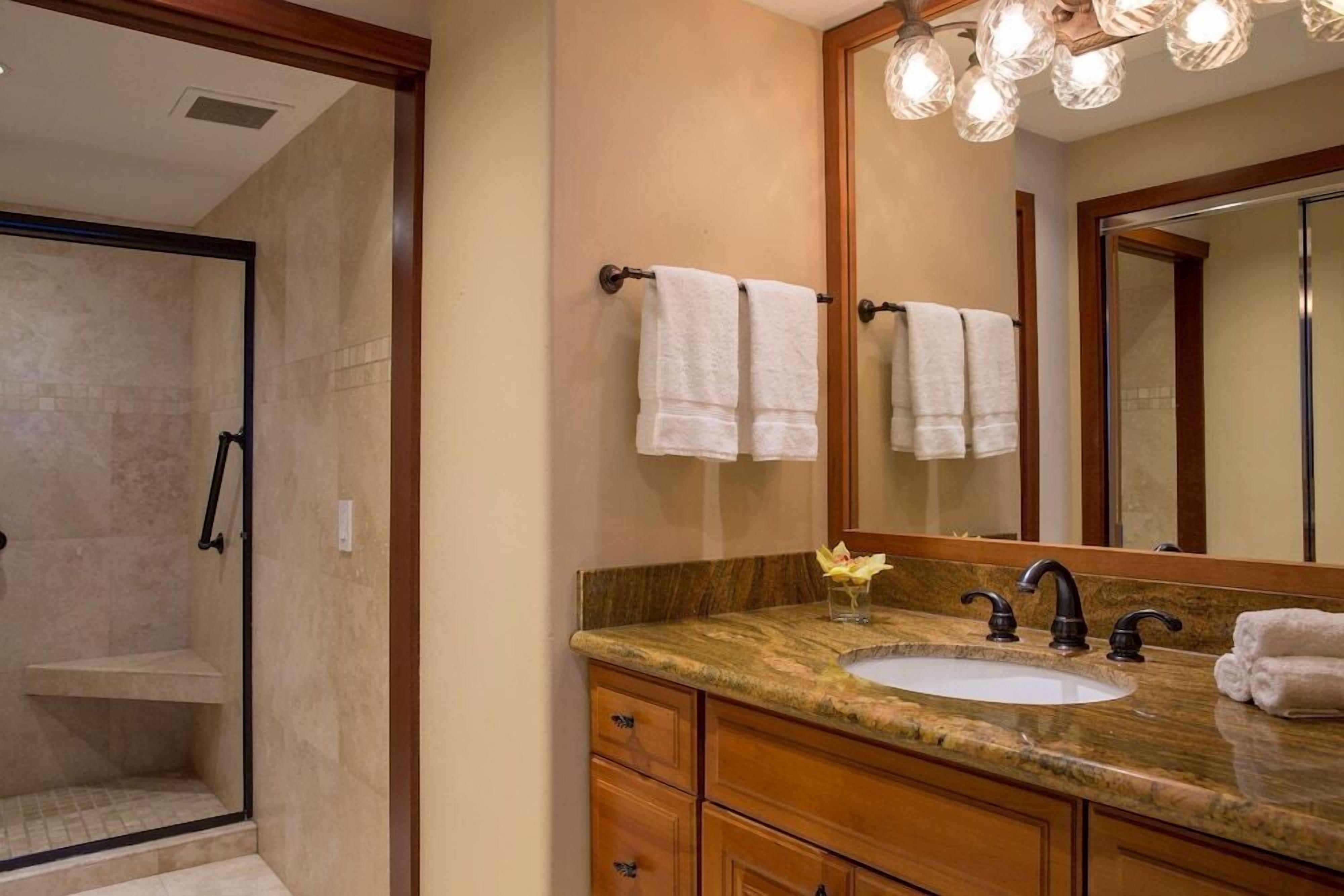A bathroom with a shower, a sink with a marble countertop, wooden cabinets, and a large mirror with a towel rack.