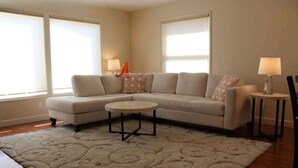 A living room with a beige sectional sofa, a round coffee table, and a patterned rug. There are two lamps on side tables and a window with blinds.