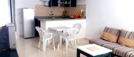 A compact living space with a kitchenette, a dining area with white chairs, a sofa with striped cushions, and a small table.