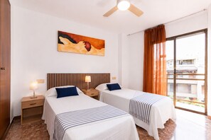 Une chambre d’hôtel avec deux lits, un ventilateur de plafond, un tableau au mur et une vue dégagée sur l’extérieur grâce à une grande fenêtre.
