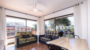 A room with a sofa, a dining table, and chairs, large windows with curtains, and a ceiling fan. - Centrally-Located Apartment · AirCon · Shared Pool · UHC CASALMAR 223 (Salou)