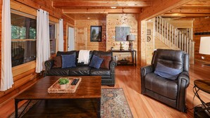 Ein gemütliches Blockhaus-Interieur mit Ledersofa, Liegesessel, Holztisch und einer Treppe, die in eine weitere Ebene führt.