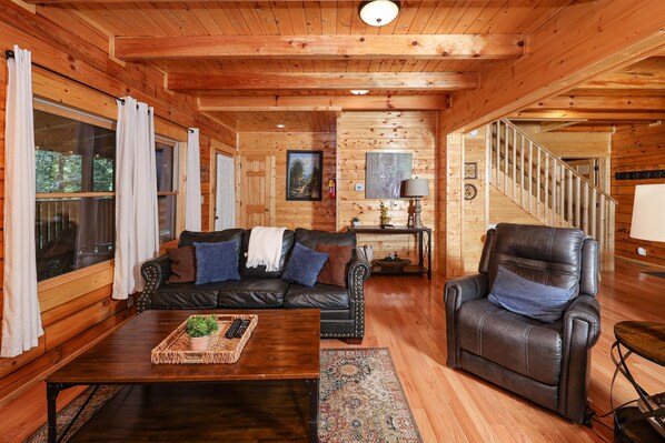 A cozy log cabin interior with a leather sofa, a recliner, a wooden coffee table, and a staircase leading to another level. - Private Fire Pit, Wheelchair Accessible, Hot Tub, Pool Table (Sevierville)