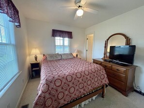 Een slaapkamer met een bed, een dressoir, een televisie, een plafondventilator, een raam met jaloezieën en een nachtkastje met een lamp.
