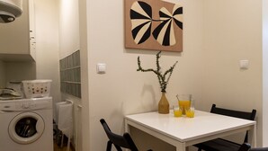 Et kompakt køkken med vaskemaskine, et spisebord med stole, en vase med grønne planter og to glas appelsinjuice.