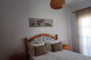 Un dormitorio con cabecera de madera, cama con ropa de cama blanca y marrón, una obra de arte colgada en la pared, una mesita de noche con lámpara y una ventana con cortinas.