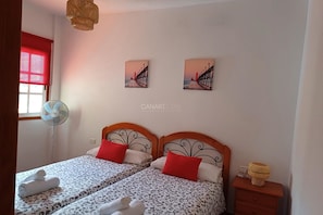 A bedroom with two beds, a standing fan, a red window blind, and two wall-mounted pictures.
