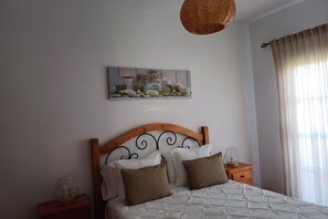 Un dormitorio con cabecera de madera, cama con ropa de cama blanca y marrón, una obra de arte colgada en la pared, una mesita de noche con lámpara y una ventana con cortinas.
