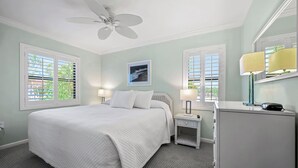 Une chambre à coucher avec un lit, deux tables de chevet avec des lampes, un ventilateur de plafond et une fenêtre avec des stores.