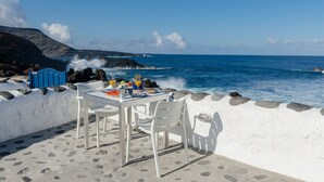 A table with chairs set on a sandy area by the sea, overlooking the ocean with rocky cliffs in the background.