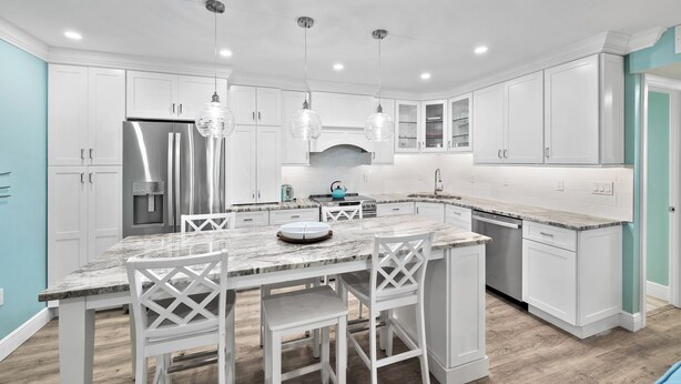 A modern kitchen with white cabinetry, a stainless steel refrigerator, a marble countertop, and a central island with bar stools.