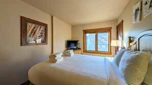 Una habitación de hotel con una cama grande, un televisor sobre una mesita de noche de madera, una ventana con vistas a edificios y un cuadro en la pared.
