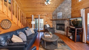 A cozy log cabin interior with a stone fireplace, leather sofa, and a television on a wooden stand. - Air Hockey, Hot Tub. FREE Amenities, Pool, Putt Putt, Fishing, Tennis, etc (Sevierville)