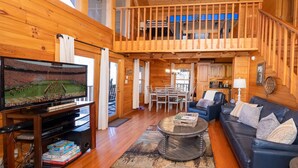 A spacious cabin with a wooden interior, a balcony, a dining area, and a living room with a sofa and a coffee table. - Air Hockey, Hot Tub. FREE Amenities, Pool, Putt Putt, Fishing, Tennis, etc (Sevierville)