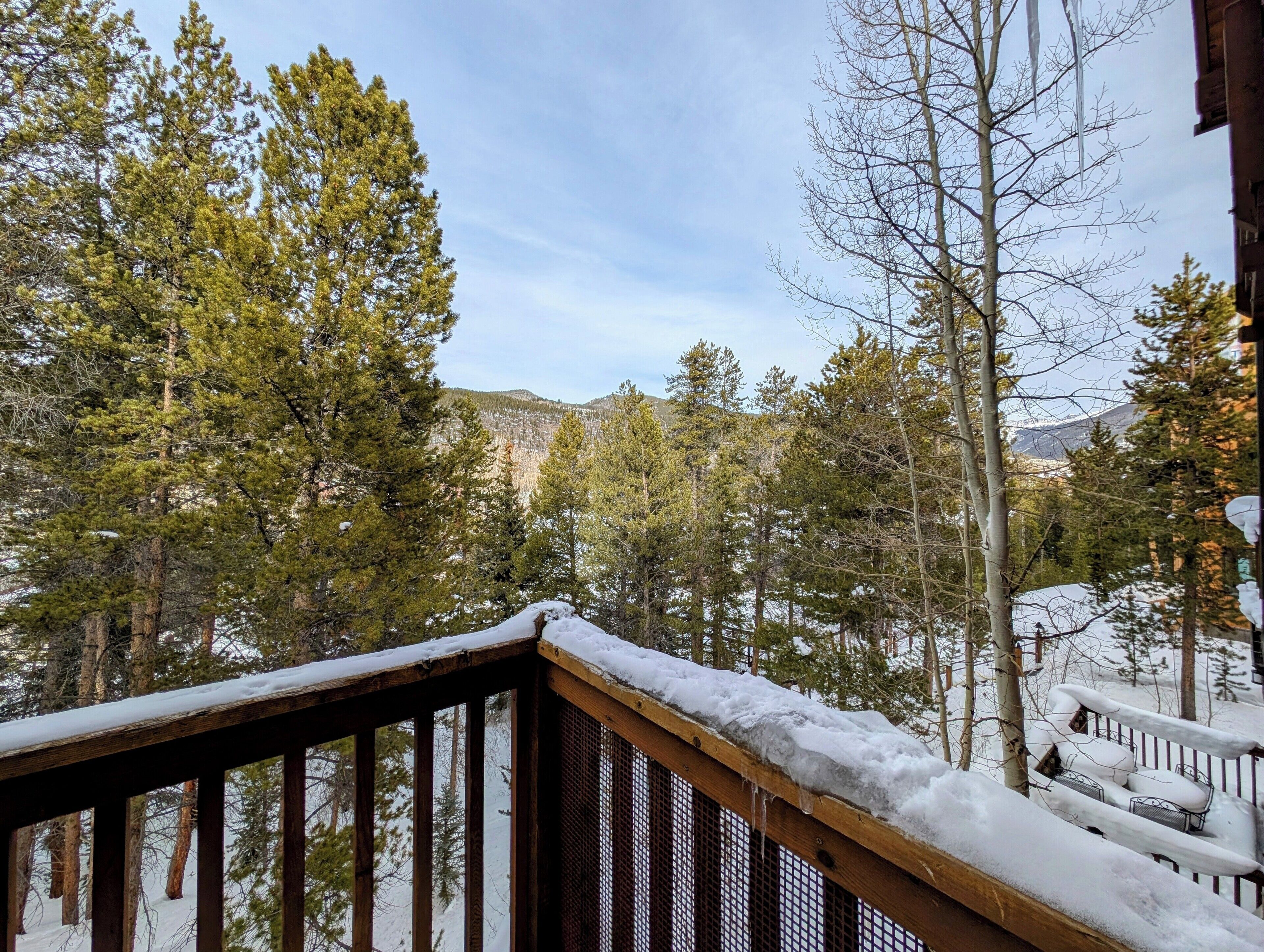 A wooden deck with snow-covered railings overlooking a forested area with trees and a distant mountain.