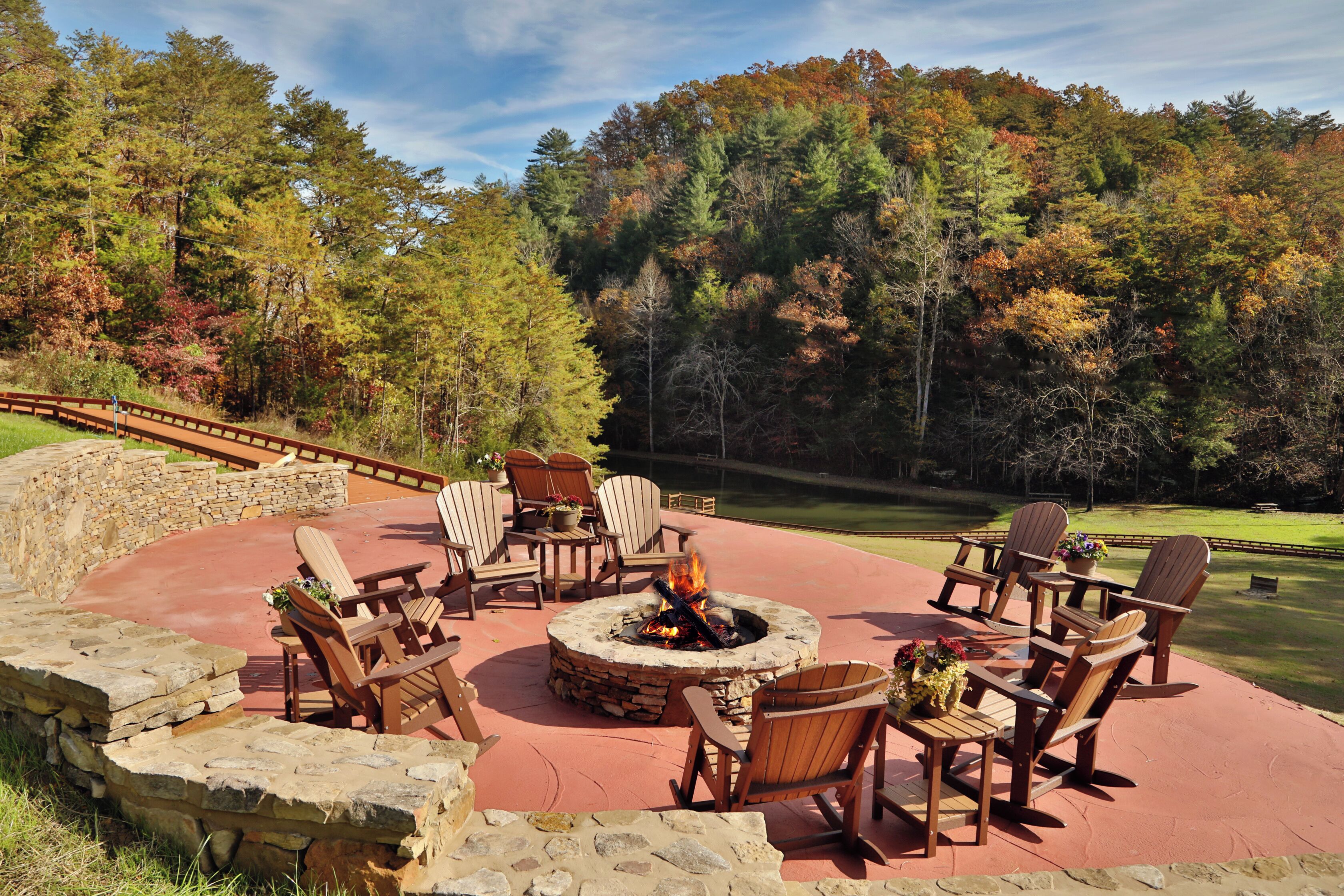 A circular stone fire pit surrounded by wooden Adirondack chairs, set on a red patio with a stone wall and a view of a golf course and forest.
