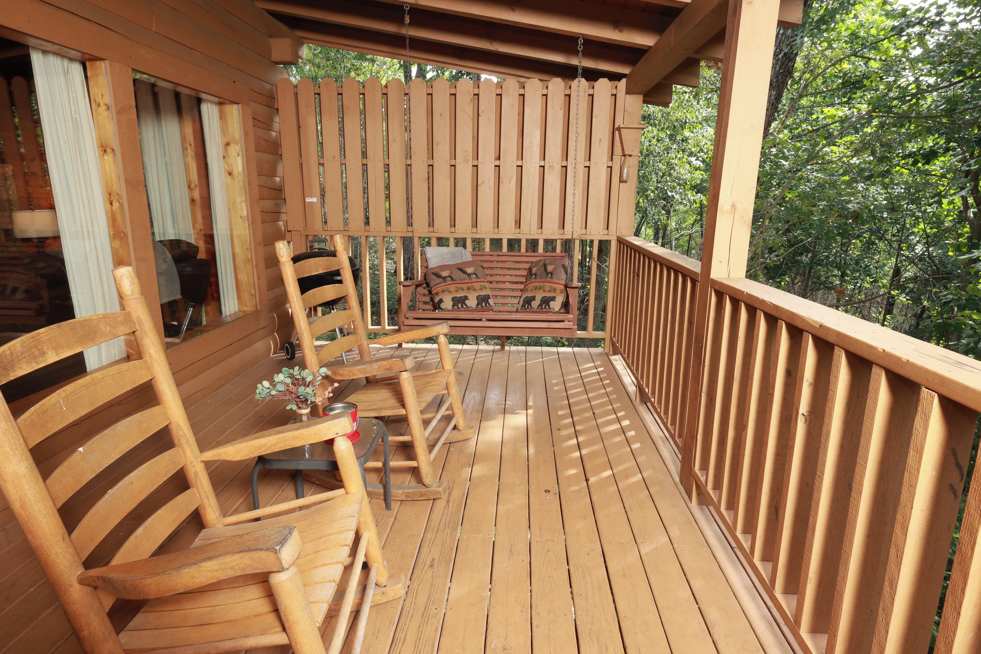 Une terrasse en bois avec un banc et des chaises en bois, une petite table avec un vase, et une vue sur les arbres.