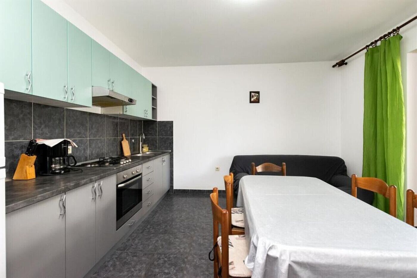 A compact kitchen with grey cabinets, a stove, and a sink. There is a dining table with chairs and a bed in the room.