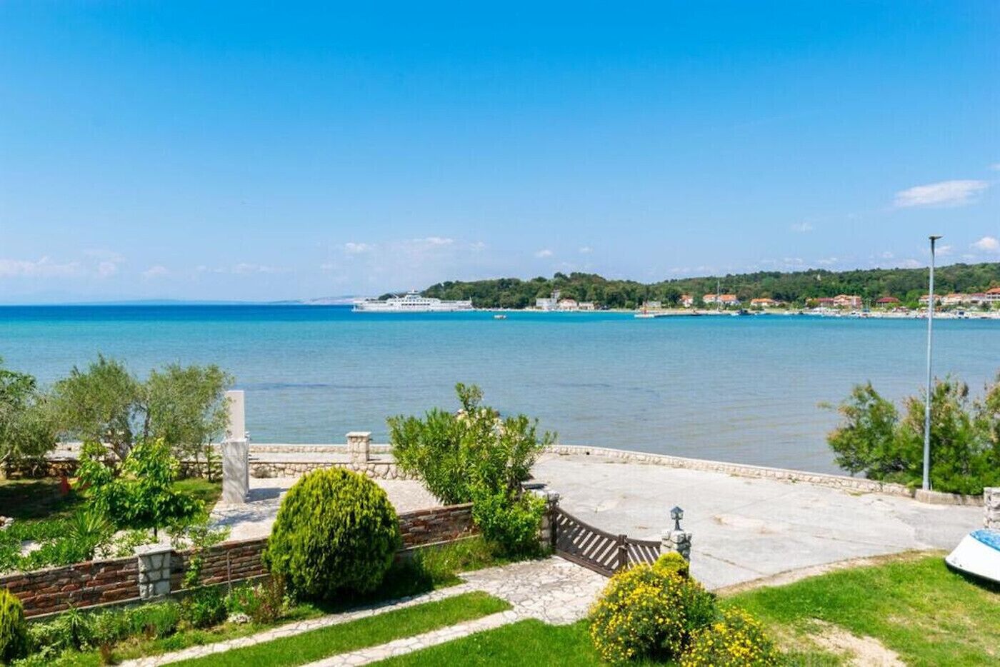 A coastal view with a clear blue sky, a calm sea, and a stone pathway leading to a grassy area with trees and a fence.