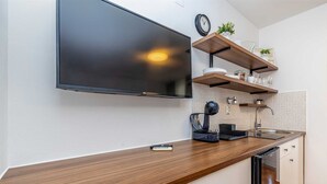 A modern kitchen with a flat-screen TV mounted above the counter, wooden shelves with kitchenware, a coffee machine, and a sink.