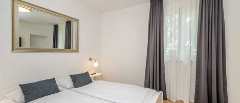 A bedroom with a bed, a mirror, a bedside lamp, and a window with curtains.
