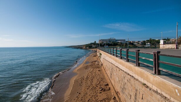 Un paseo marítimo con una playa arenosa, una barrera de hormigón y edificios al fondo.
