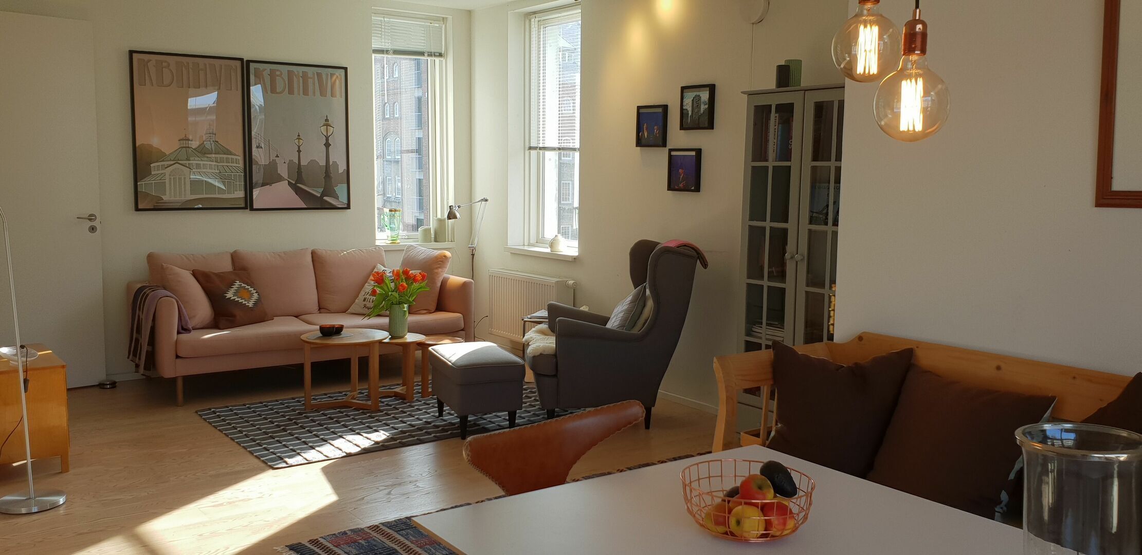 A cozy living room with a sofa, armchair, and coffee table. There are framed pictures on the wall and a window with blinds.