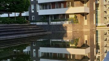 A multi-story residential building with balconies, a reflective water body in front, and a clear sky.