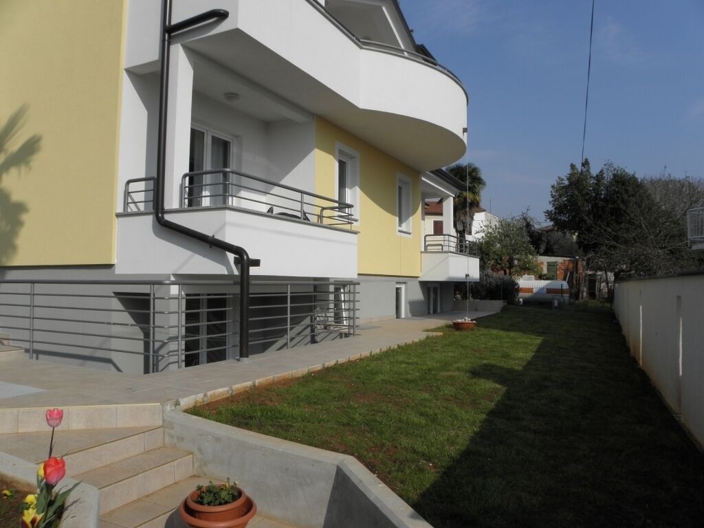A modern two-story building with a flat roof, a balcony, and a small garden area with steps leading to the entrance.