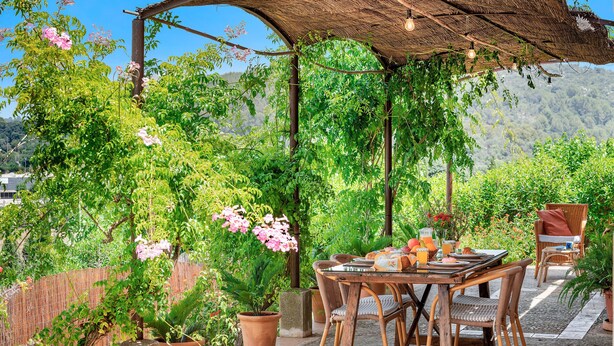 En udendørs terrasse med et træbord og fletstole, omgivet af plantede bede og et stråtækt tag.