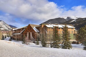 A lodge with a wooden exterior and multiple stories, surrounded by snow-covered mountains and trees.