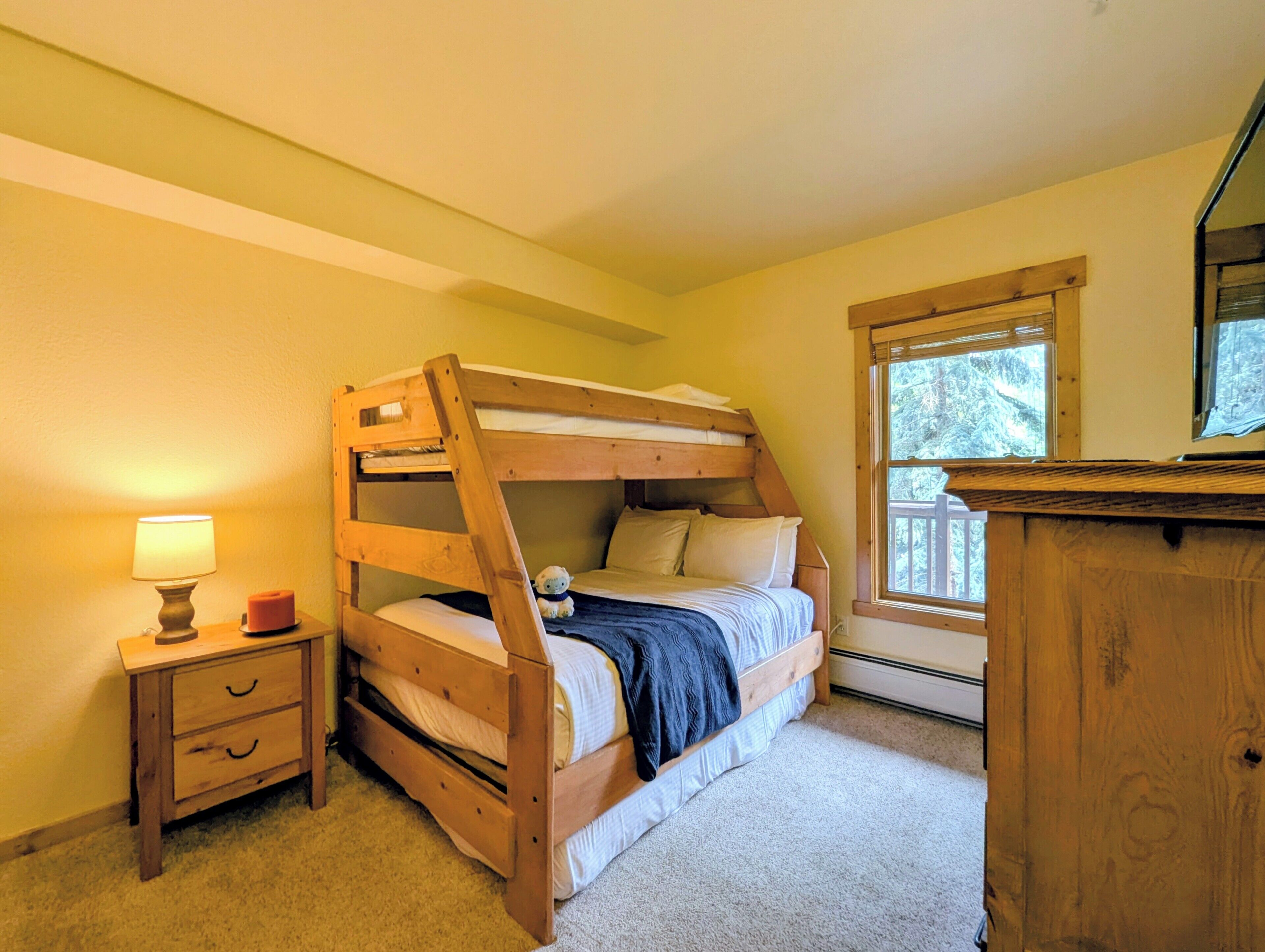 Una cama individual con cabecera de madera, una mesita de noche con lámpara, un aparador y una ventana con vista a árboles.