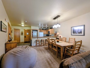 A compact kitchen with wooden cabinets, a refrigerator, and a microwave. There is a dining area with a table and chairs, and a large bean bag chair.
