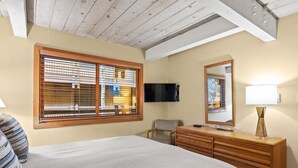 Een slaapkamer met een bed, een houten dressoir, een stoel, een televisie en een raam met jaloezieën.