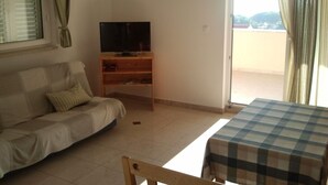 Una sala de estar con un sofá, un televisor sobre un mueble de madera, una mesa con mantel a cuadros y vistas a través de un ventanal.