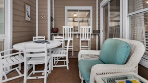 A covered patio area with a round table and white chairs, a wicker chair with a teal cushion, and a window with blinds. - Updated Unit! Pictures coming Soon! (Miramar Beach)