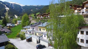 Un village perché sur une colline, avec ses maisons traditionnelles en bois, une voiture garée devant, et une chaîne de montagnes en arrière-plan.