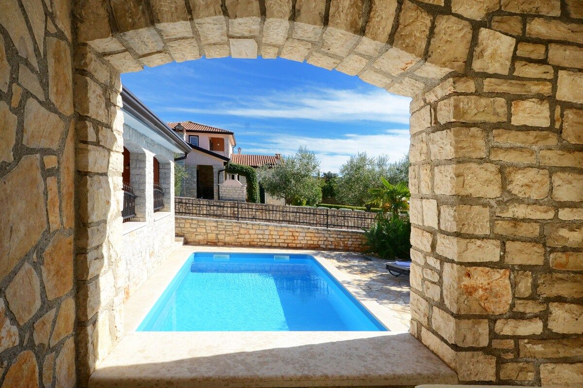 A stone archway leading to a swimming pool with a house in the background.