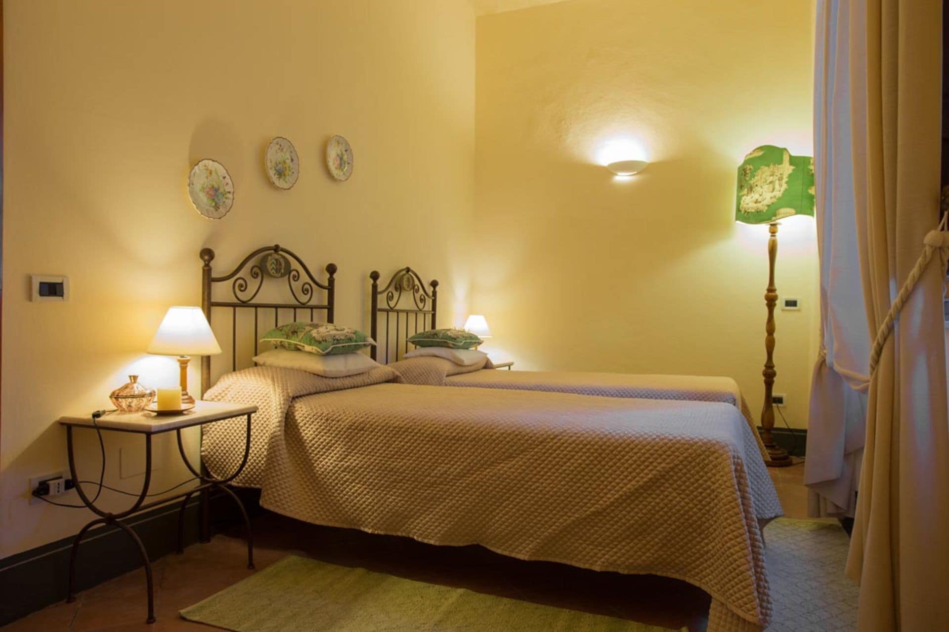 A hotel room with two single beds, a bedside table with a lamp, a green floor lamp, and a small table with a candle.