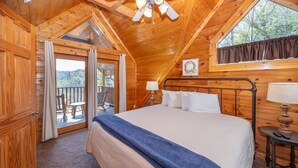 A wooden cabin bedroom with a large bed, a nightstand, a window with curtains, and a balcony door leading outside. - Great Mountain Views! 5miles to Pigeon Forge - Pool, Fishing, Putt Putt, Golf (Sevierville)