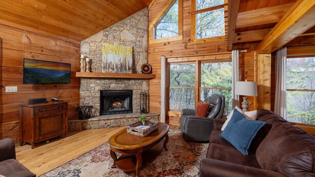 A cozy cabin interior with a stone fireplace, wooden walls, a large window, and a leather sofa. - Great Mountain Views! 5miles to Pigeon Forge - Pool, Fishing, Putt Putt, Golf (Sevierville)
