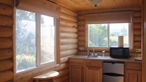 A log cabin kitchen with wooden cabinets, a sink, a microwave, and a window providing a view of the outdoors. - Wranglers Quarters RiverView Cabin - Sevier River Ranch - 1 King bed (Hatch)