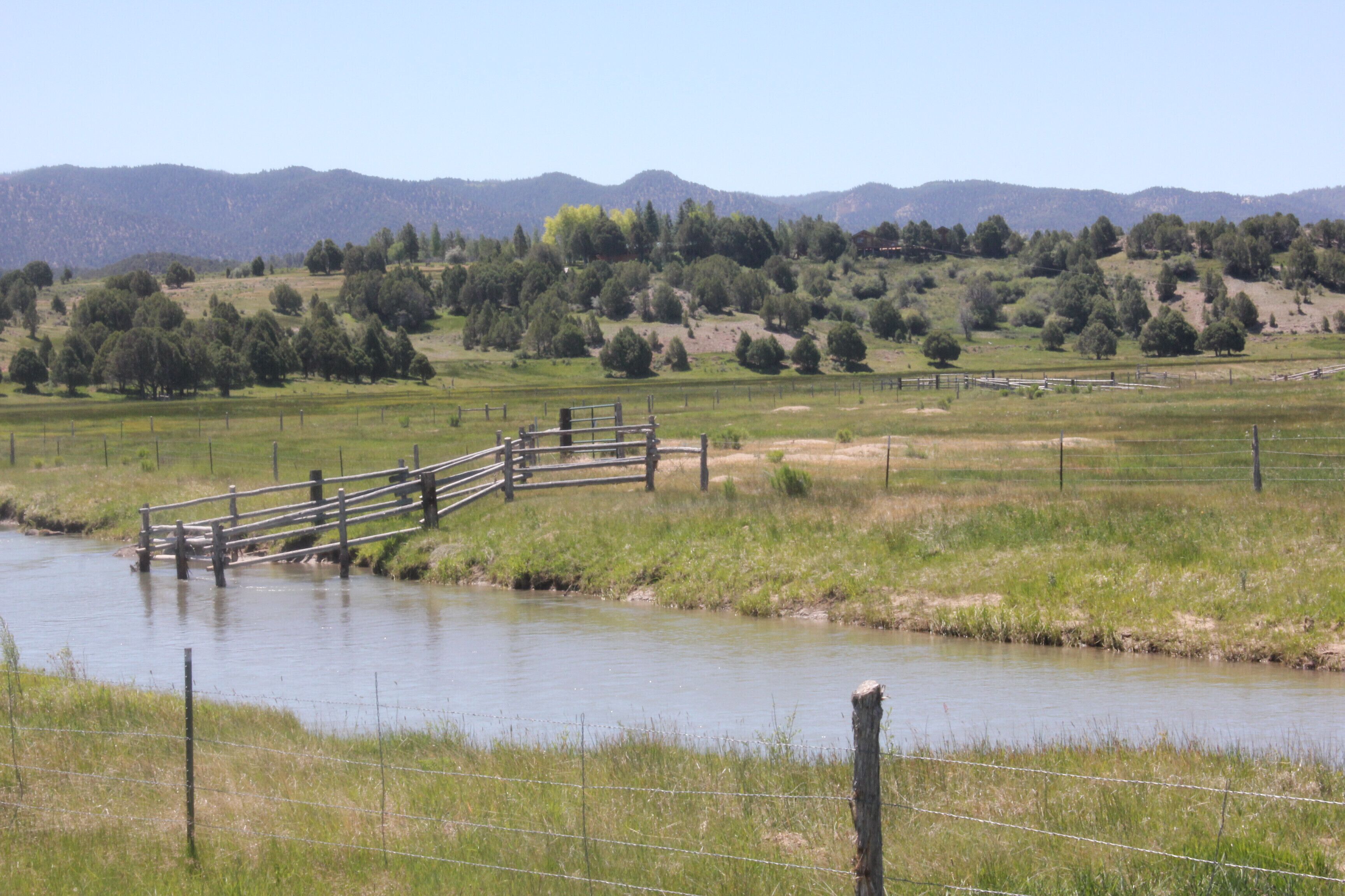 Un pont en bois enjambe une rivière paisible, entouré de prairies verdoyantes et d’arbres, avec des montagnes en arrière-plan.
