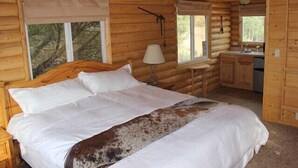 A log cabin bedroom with a wooden bed, a nightstand, a desk, and a window with a view of trees. - Wranglers Quarters RiverView Cabin - Sevier River Ranch - 1 King bed (Hatch)
