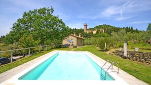 Ein Schwimmbad mit einem Steinhaus im Hintergrund, umgeben von viel Grün und einem Hügel mit einem Turm.