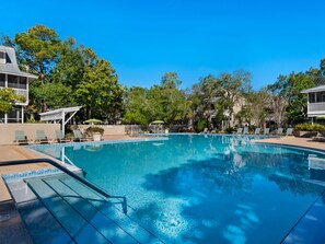Ein klares, blaues Schwimmbecken, umgeben von Liegestühlen und Sonnenschirmen, mit Bäumen und Gebäuden im Hintergrund.