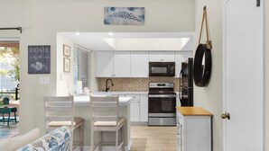 Een moderne keuken met witte kastjes, een magnetron, oven en een koelkast. Er is een eethoek met een tafel en stoelen.
