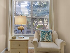 A room with a window, a lamp on a nightstand, a chair with a cushion, and a small table with a bowl and a device.