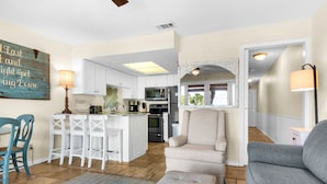 Amplia sala de estar con un sofá gris, un sillón beige y un reposapiés blanco. Se ve una cocina con armarios blancos y una isla central al fondo.