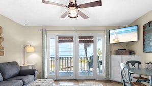 Una sala de estar con un sofá gris, un taburete capitonado, una mesa de comedor con sillas azules, un ventilador de techo y un televisor que muestra una escena de playa.